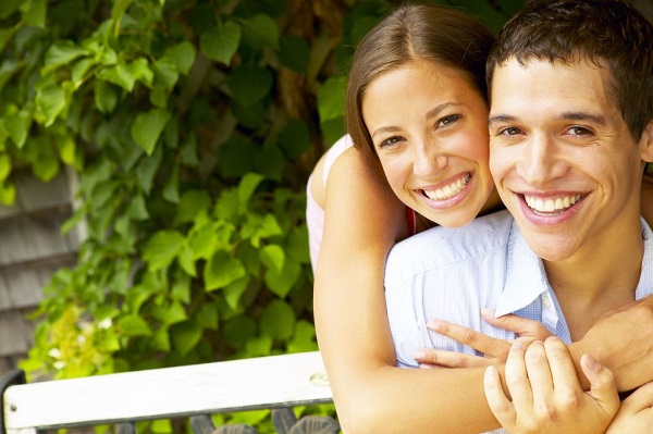 Headshot of a young man in collared shirt smiling with a girl hugging him from behind and smiling with nature background
