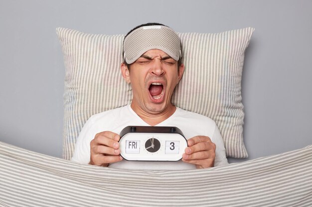 funny-man-with-resting-white-pillow-blanket-wearing-tshirt-isolated-grey-background-holding-alarm-yawning-with-widely-open-mouth-grimacing-face